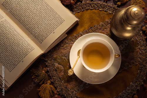 Toma cenital de taza de té de porcelana con cucharita dorada, mantel bordado y lectura de acompañamiento  photo