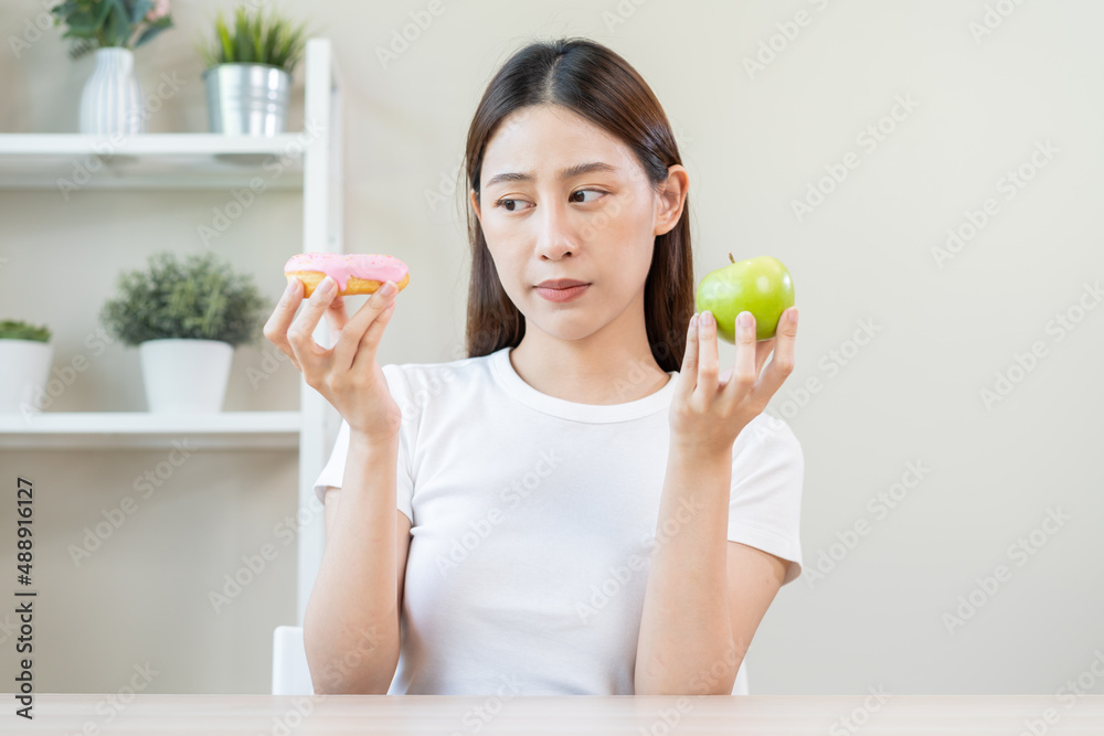 Diet, Dieting asian young woman or girl smile, confused choose, choice green apple or pink sweet donut, at home, eat food for good healthy, health when hungry. Close up female weight loss person. Diet, Dieting asian young woman or girl smile, confused choose, choice green apple or pink sweet donut, at home, eat food for good healthy, health when hungry. Close up female weight loss person.
