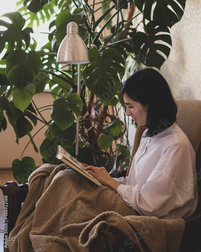 Принти на полотні Young woman is reading a book and sitting at home in a chair with bedcover