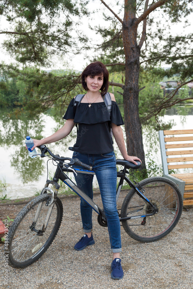 Obraz premium An adult brunette woman in jeans and a blouse with a backpack rides a bike in a park by the river.