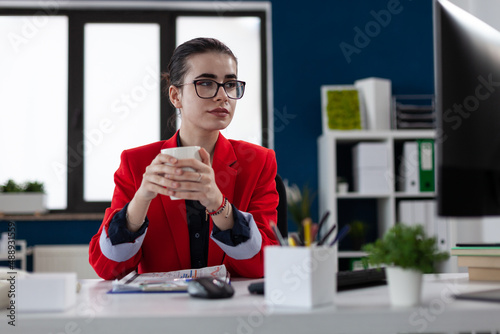 Small business owner with glasses at startup desk holding white cup. Employee in red jacket having coffe or tea break and loking at compouter screen. Businesswoman taking a pause from working.