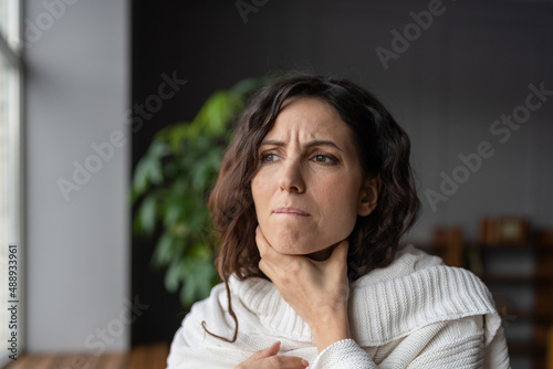 Young sick worried woman with sore throat at home touching neck, unhealthy female wrapped in plaid having swollen lymph nodes caused by bacterial infection, feeling pain or discomfort while swallowing