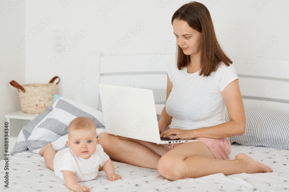 Indoor shot of dark haired woman wearing white shirt and short sitting on bed with her daughter and working on laptop, combines maternity leave and online job.