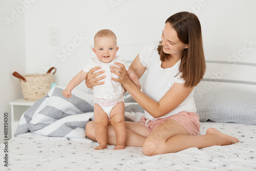 Portrait of positive satisfied woman wearing white shirt and short sitting on bed with her daughter in body suit, mother holding her standing toddler child, dark haired female playing with kid.