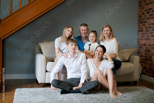 Portrait caucasian happy big family smiling at camera in cozy living room. Joyful man and woman sitting on couch in an embrace with children and granddaughter. Parents and children at home generations