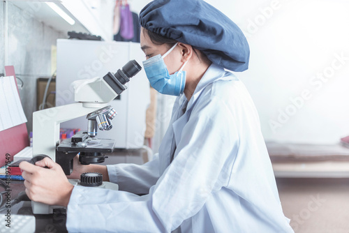 An asian laboratory technician checks a swab specimen sample with an optical microscope. Working at a research facility of hospital lab. Wearing a bouffant cap and face mask. photo