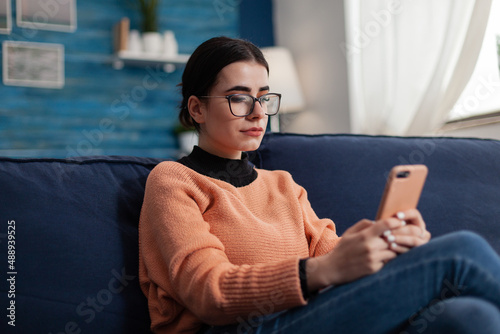 Student relaxing on couch holding smartphone and looking at screen. Content creator with glasses on sofa reading content on mobile phone. Digital influencer checking email on tochscreen device. photo