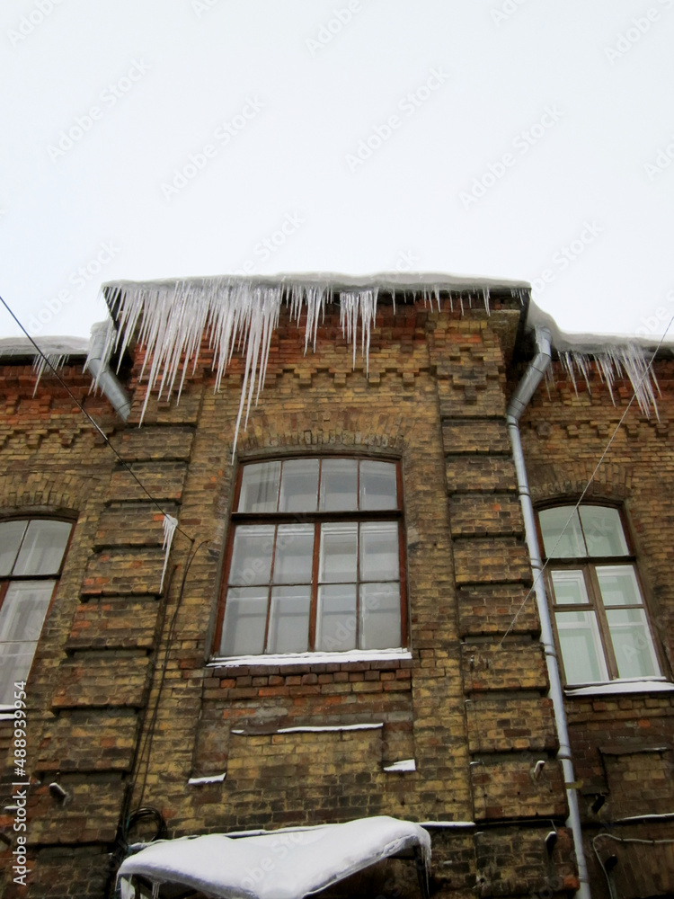 Huge icicles of ice on the roof of an old house. Vertical photo with icicles on the roof. Poor ice cleaning. Danger on the roof of the house. Huge icicles of ice on the roof of an old house. Vertical photo with icicles on the roof. Poor ice cleaning. Danger on the roof of the house.