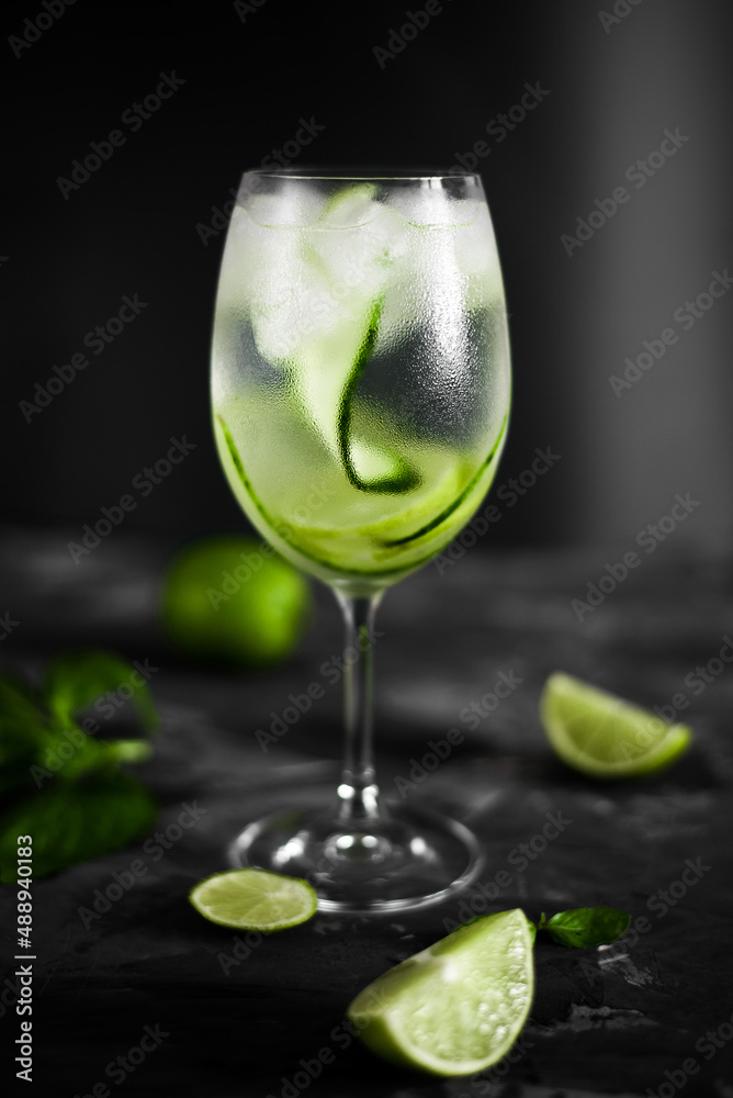 A wine glass of fresh cold coctail with ice, cucumber slices, mint and lime fruit on  the dark background