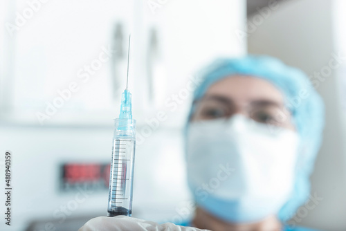 An asian nurse in ppe gear checks a syringe filled with drugs prior to injection. Selective focus on the needle. Hospital or ER scene.