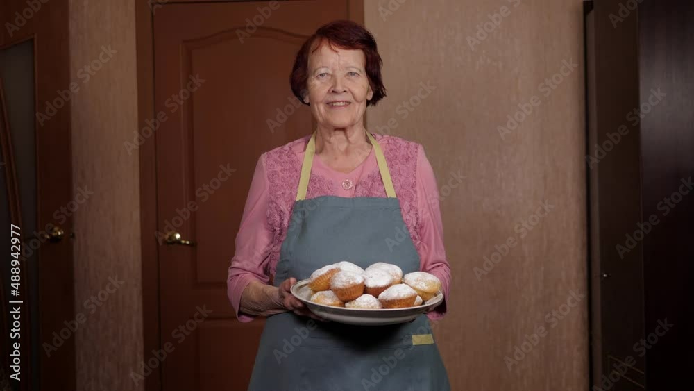 Portrait of a caring grandmother in an apron with a tray of cupcakes in her hands, she stands at home and looks into the camera. Portrait of a caring grandmother in an apron with a tray of cupcakes in her hands, she stands at home and looks into the camera.