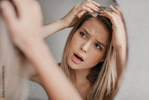 Frustrated woman searching hair flakes suffering from dandruff problem, looking at her reflection in mirror in bathroom