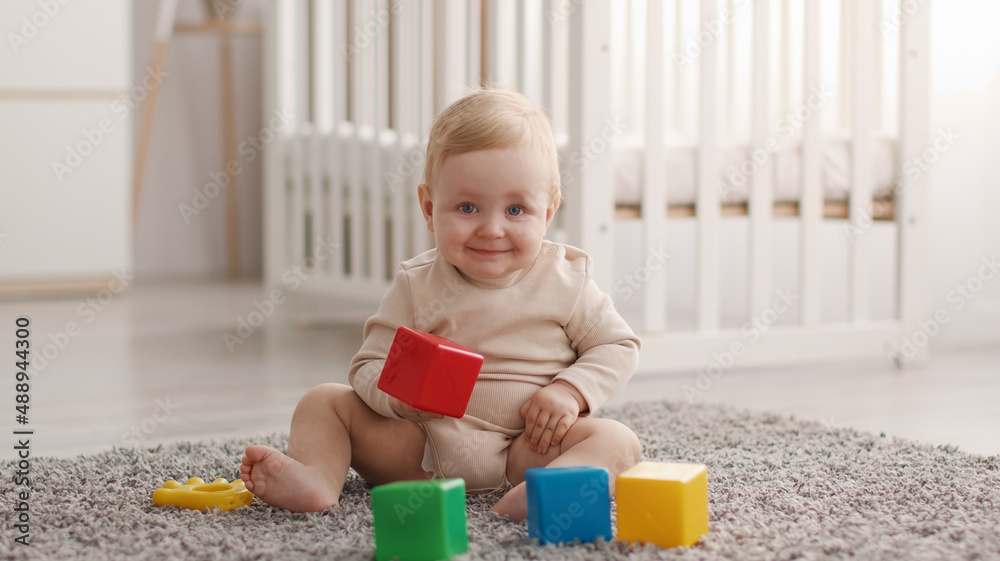 Portrait of adorable little baby smiling to camera, playing with colorful cubes, resting on floor at nursery, panorama