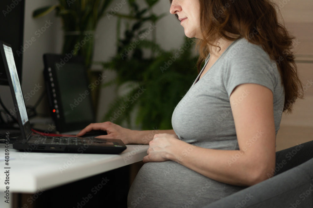 A pregnant woman is working on a laptop. The hand is on the stomach. Pregnant businesswoman typing on a laptop while sitting at her workplace in the office. Work during pregnancy from home.