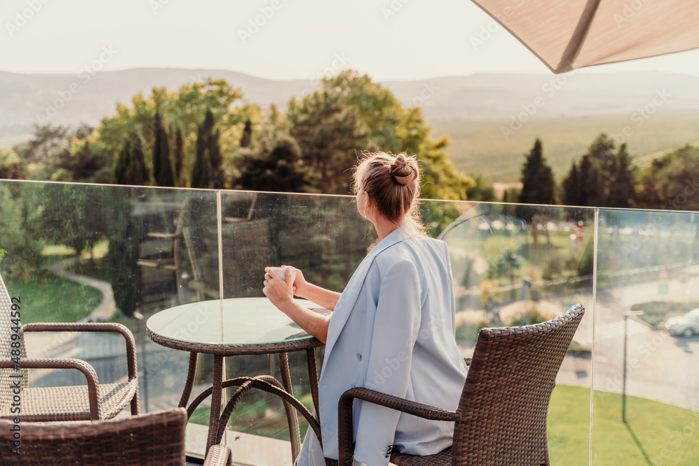 A middle-aged woman sits in a street cafe overlooking the mountains at sunset. She is dressed in a blue jacket and drinks coffee admiring the nature. Travel and vacation concept. A middle-aged woman sits in a street cafe overlooking the mountains at sunset. She is dressed in a blue jacket and drinks coffee admiring the nature. Travel and vacation concept.