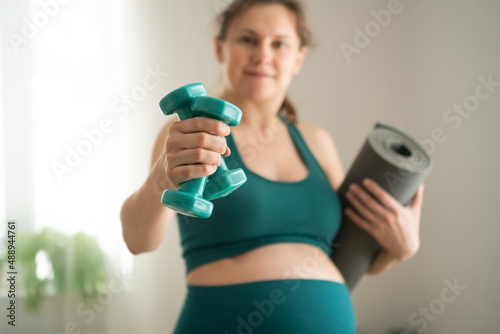 A pregnant woman prepare for exercising with dumbbells. Young girl holding a yoga mat in hand. Concept of a healthy lifestyle during pregnancy. Sports activities before the birth of baby.