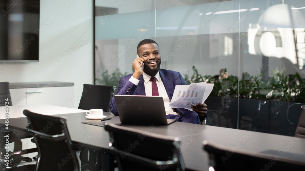 Smiling black boss having phone conversation at office, holding documents Smiling black boss having phone conversation at office, holding documents