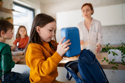 Little girl packing lunch box to backpack in kitchen at home.