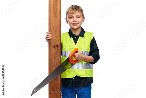 building, carpentry and profession concept - happy smiling little boy in protective helmet and safety vest with saw and wooden board over white background