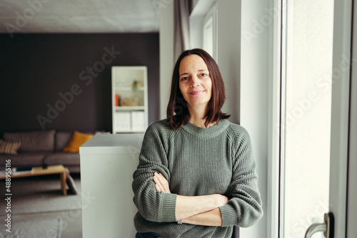 Portrait of smiling woman with arms crossed leaning by window at home