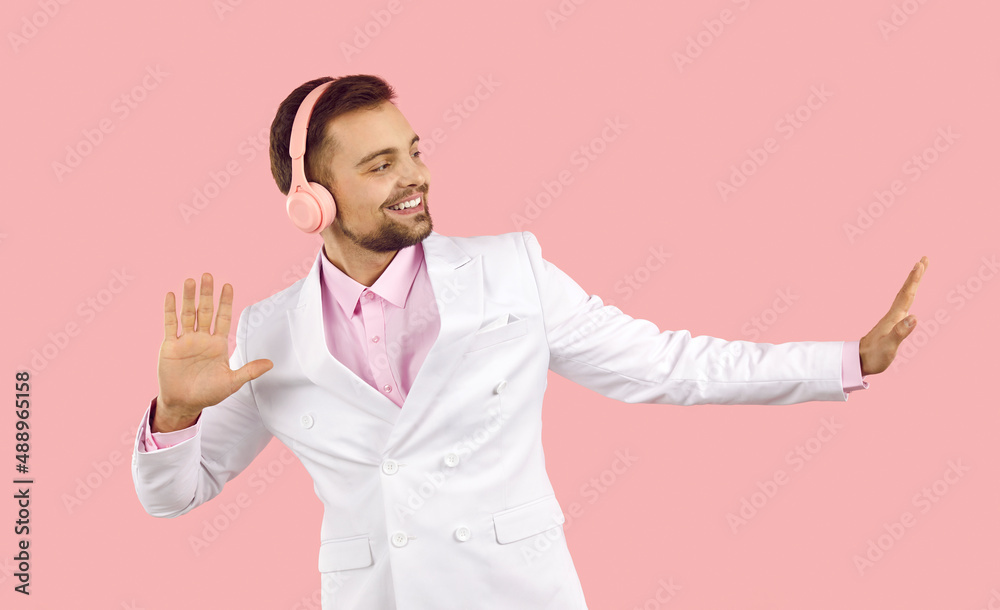 Happy cheerful relaxed handsome young man wearing elegant white suit, pink shirt and modern headphones listening to his favourite music and dancing isolated on solid pastel pink colour background Happy cheerful relaxed handsome young man wearing elegant white suit, pink shirt and modern headphones listening to his favourite music and dancing isolated on solid pastel pink colour background