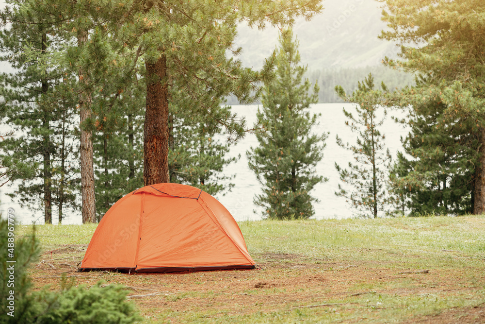 A lonely orange tent is set up on soft green grass in the forest near the lake. Weekend activity and hiking or trekking adventures