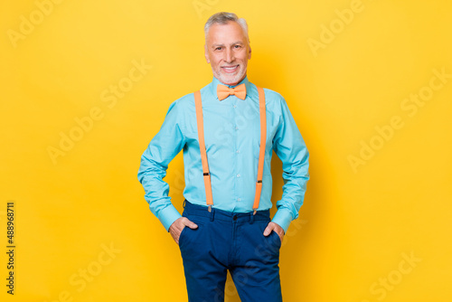 Photo of elder grey hairdo man wear teal shirt suspenders bow tie trousers isolated on yellow color background