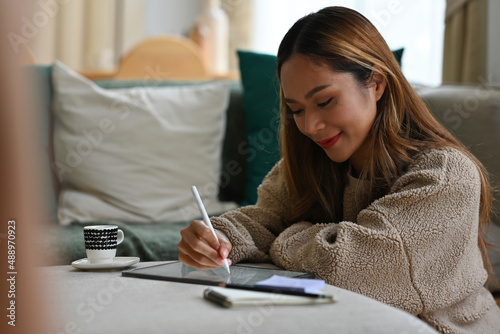 Side view portrait of a young pretty Asian woman sitting on a floor working on tablet in a modern living room  for work and technology concept.