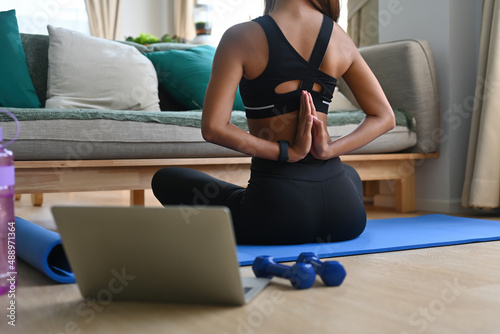 Fototapeta Naklejka Na Ścianę i Meble -  A cropped view portrait of a young  woman stretching hands behind the back on a mat in a living room with dumb bells and laptop on the side, for home exercise, online class concept.