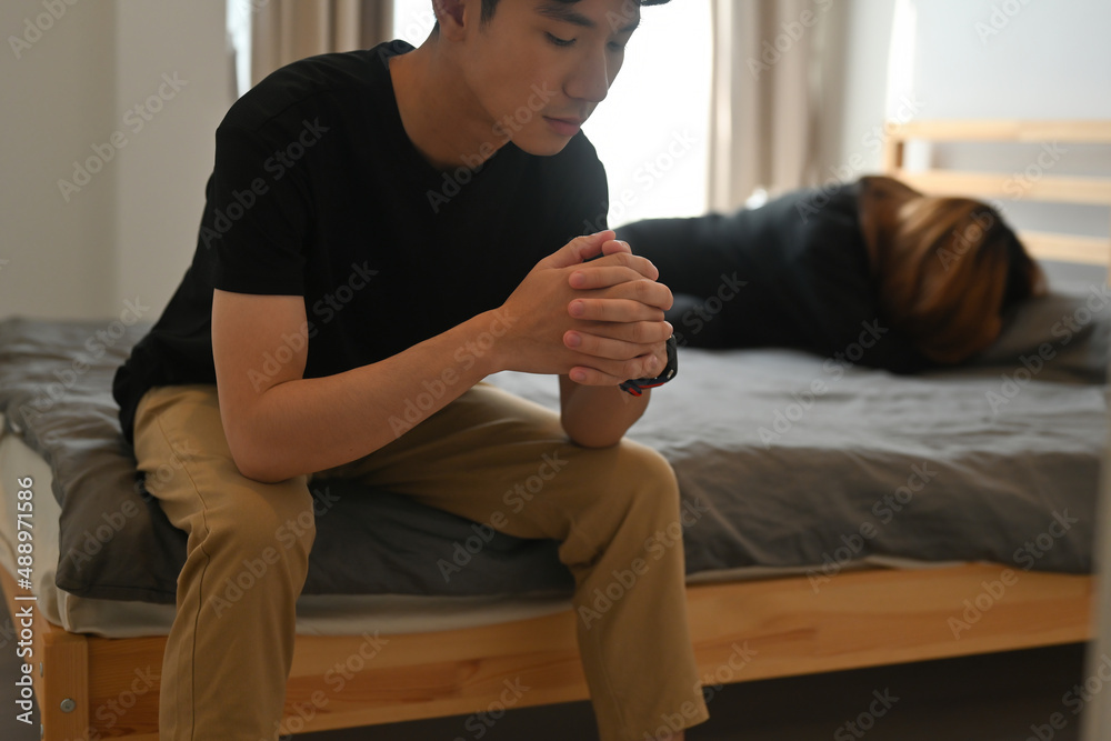 A cropped view portrait of a man sitting on a bed with a stressful gesture and a woman sleeping behind, for marriage life relationship and family life concept. A cropped view portrait of a man sitting on a bed with a stressful gesture and a woman sleeping behind, for marriage life relationship and family life concept.