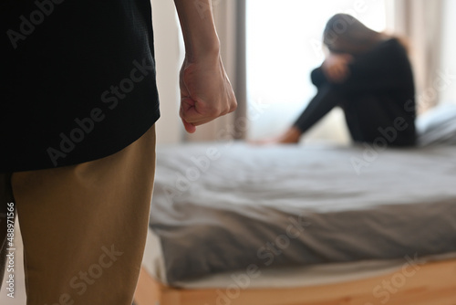 Cropped view a woman sitting on the bed with stress gesture in the background, the man's fist in the foreground. For marriage life, relationship and violence concept.