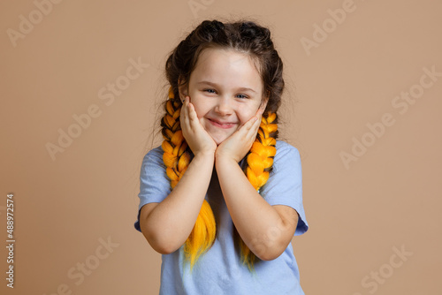 Happy, little girl touching cheeks with hands smiling looking at camera with yellow kanekalon braids on head on beige background wearing blue t-shirt and jeans.