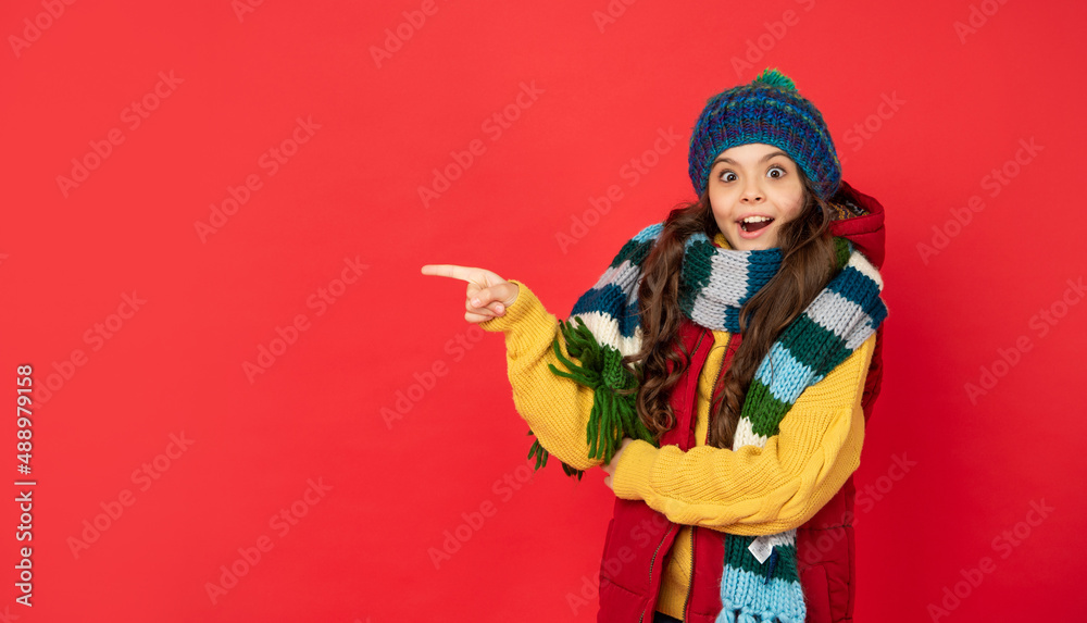 amazed kid in knitted winter hat and scarf pointing finger on copy space, advertising