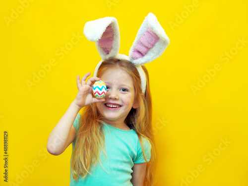 Closeup portrait of a little girl on a yellow background with an easter colored egg. Cute happy cheerful beautiful charming child in a bunny costume.