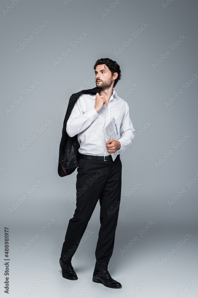 full length view of businessman holding blazer and newspaper while looking away on grey full length view of businessman holding blazer and newspaper while looking away on grey