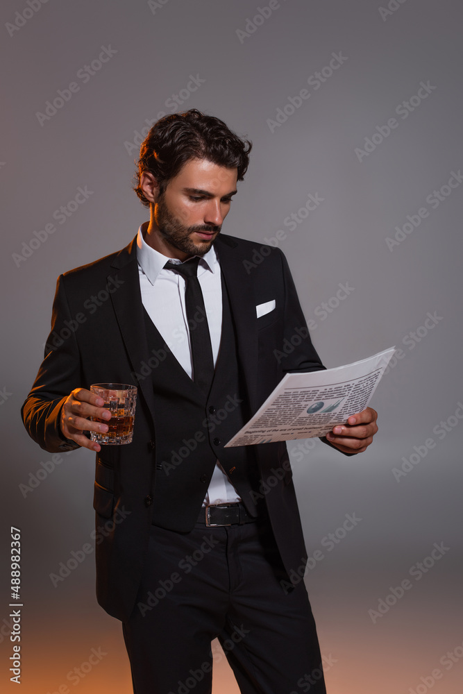 man in black elegant suit reading newspaper while holding glass of whiskey on grey background man in black elegant suit reading newspaper while holding glass of whiskey on grey background