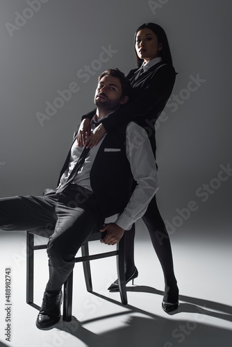 stylish man sitting on chair near sexy asian woman seducing him on grey background