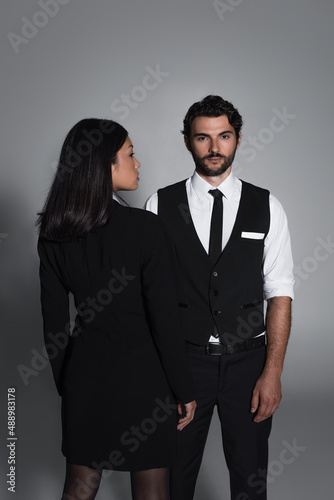 back view of asian woman in black blazer dress near stylish man looking at camera on grey background