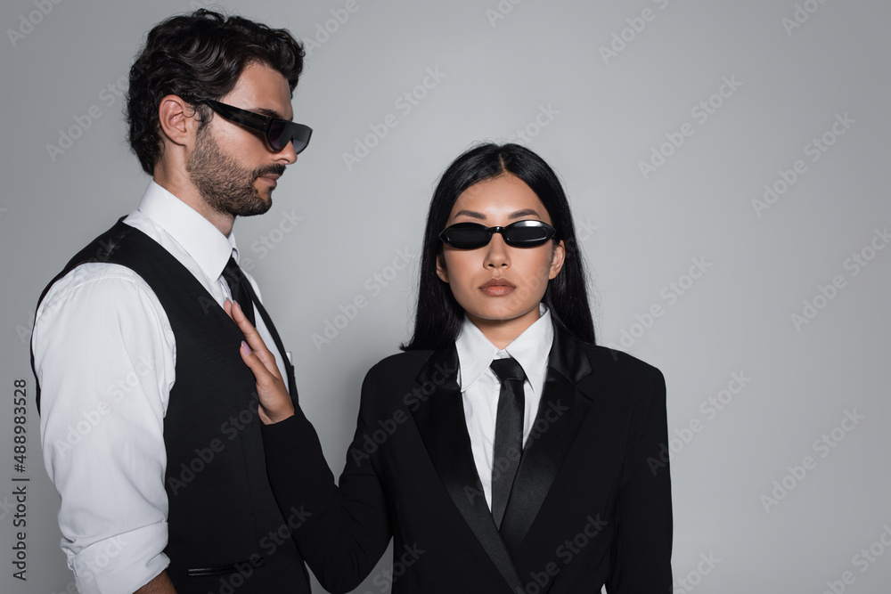 elegant asian woman in black formal wear and sunglasses looking at camera near stylish man isolated on grey elegant asian woman in black formal wear and sunglasses looking at camera near stylish man isolated on grey