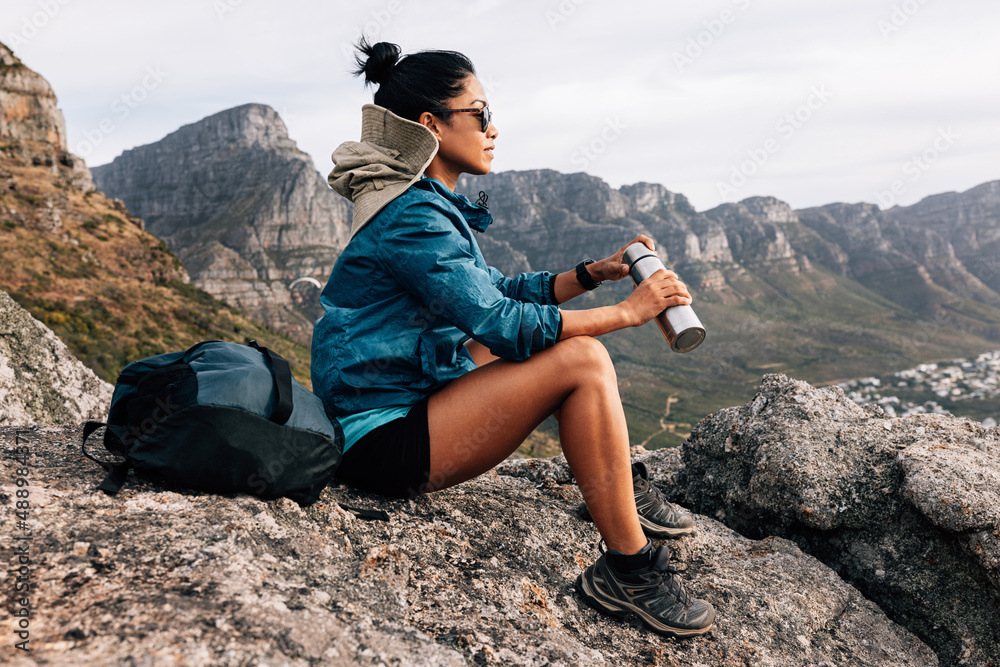 Side view of woman hiker in sports clothes enjoying the view. Female in sunglasses holding a thermos sitting on a rock and relaxing during a hike.