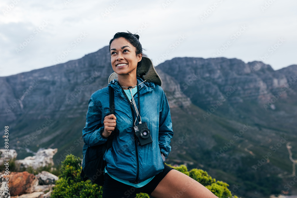 Portrait of a smiling woman relaxing during a mountain hike. Female with analog camera and backpack enjoying the view while standing on the top of the mountain.