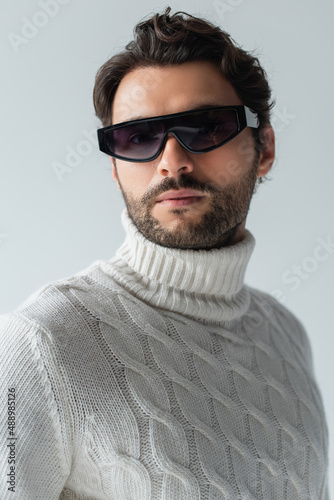 trendy man in white sweater and dark sunglasses looking at camera isolated on grey © LIGHTFIELD STUDIOS