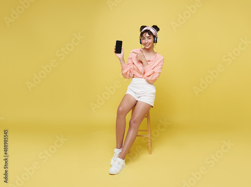 Young asian teenage girl hand pointing smart phone. While her listening music in headphones and sitting on chair isolate on yellow background.
