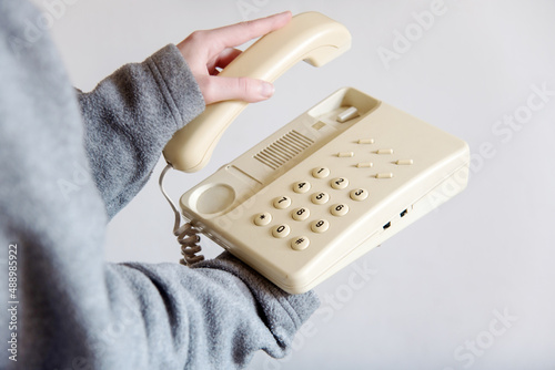Detalle de una mano de mujer descolgando un teléfono blanco antiguo con cable. Mano sosteniendo el auricular de un teléfono fijo de casa u oficina. Concepto de atender una llamada telefónica. photo