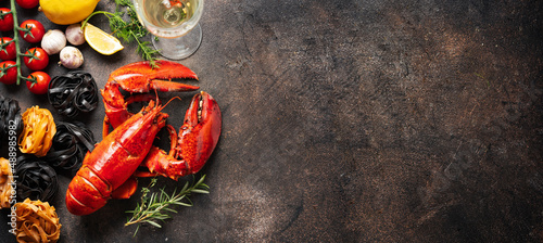 Cooked red lobters with ingredients for pasta and glass of whhite wine on dark background. Gourmet holidays food. Long banner with copy space