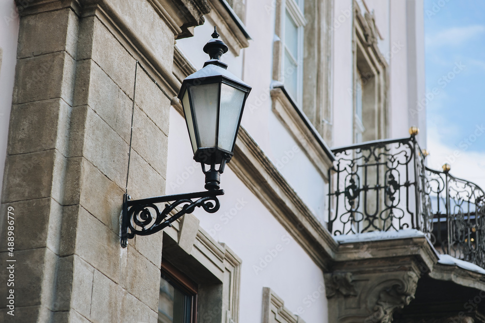Old-fashioned street wall lamp made of black metal with snow. Gray facade of an old house with windows. Winter Lviv, Ukraine. Old-fashioned street wall lamp made of black metal with snow. Gray facade of an old house with windows. Winter Lviv, Ukraine.
