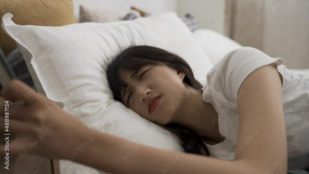 closeup view sleepy asian girl reluctantly waking up from alarm calling on phone in early morning and returning to sleep after turning it off on bed in bedroom closeup view sleepy asian girl reluctantly waking up from alarm calling on phone in early morning and returning to sleep after turning it off on bed in bedroom