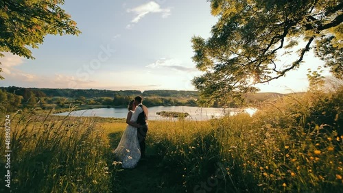 Side view of attractive bride and groom standing, hugging on field in setting sun rays. Camera approaching to wedding couple while they kissing with beautiful lake on background. Concept of wedding photo