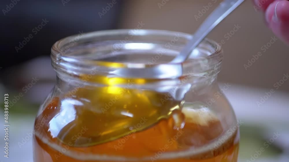 A Female Hand Picks Up with a Spoon Transparent, Liquid Honey from a Glass Jar. Yellow thick flow bee honey pouring, falling smoothly stream in tank. Healing, organic beekeeping product. Zoom. A Female Hand Picks Up with a Spoon Transparent, Liquid Honey from a Glass Jar. Yellow thick flow bee honey pouring, falling smoothly stream in tank. Healing, organic beekeeping product. Zoom.