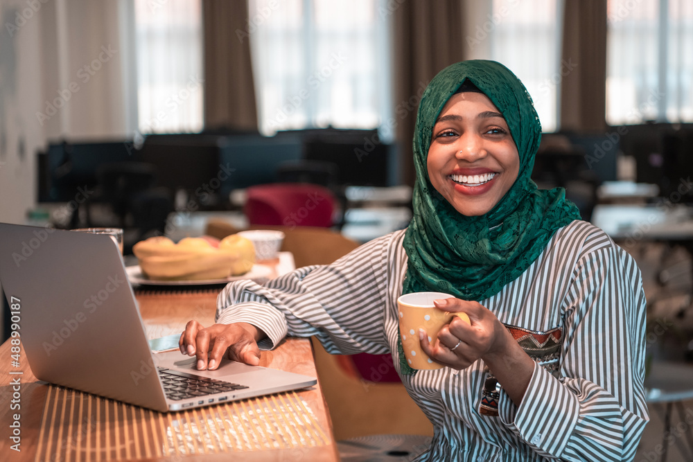 African muslim business woman wearing a green hijab drinking tea while working on laptop computer in relaxation area at modern open plan startup office. African muslim business woman wearing a green hijab drinking tea while working on laptop computer in relaxation area at modern open plan startup office.